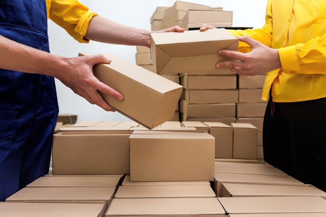 Två lagerarbetare som hanterar kartonger i ett e-handelslager, en process som tydligt visar paketflödet och som kan optimeras avsevärt med automation för att säkerställa snabb och korrekt orderhantering.